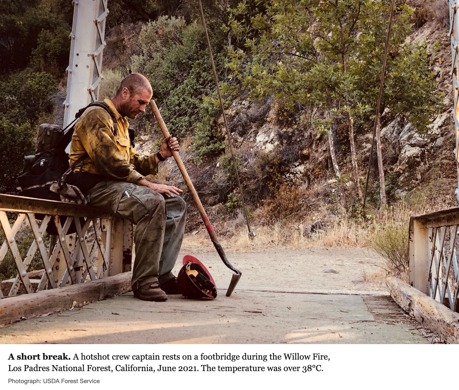 A news photograph: a wildland firefighter sits on the rail of an old metal footbridge at the left of the frame, a pack on his back. His yellow shirt is smeared with soot. He leans his head against the long wooden handle of his tool, eyes down, one hand resting on his knee; his red helmet lies on the ground by his boots. The rest of the frame is the empty bridge and a dry, steep, scrubby hillside in warm sunlight. Caption: ‘A short break. A hotshot crew captain rests on a footbridge during the Willow Fire, Los Padres National Forest, California, June 2021. The temperature was over 38°C.’ Credit: ‘Photograph: USDA Forest Service’.