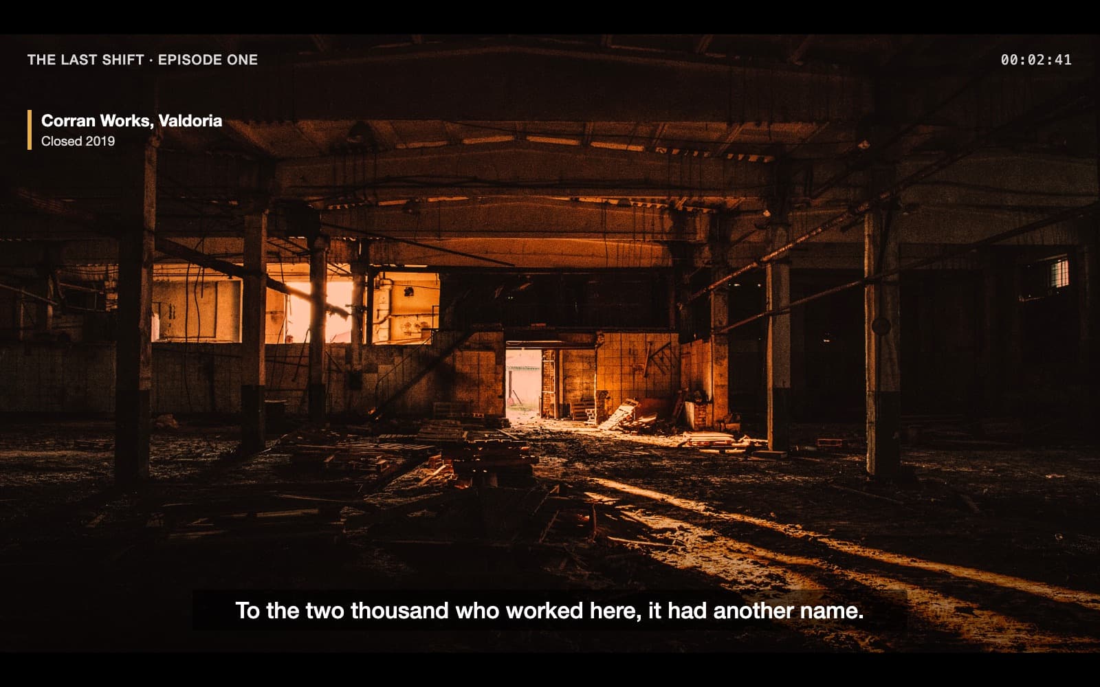 A 16:9 frame from a documentary: a vast, dark, empty factory hall. Rows of bare concrete pillars hold up the roof beams; cables hang loose; broken pallets and rubble litter the floor. Far away, at the back, daylight pours through one open doorway and falls in long stripes across the floor. Top left: ‘THE LAST SHIFT · EPISODE ONE’; top right the timecode 00:02:41; below, a place caption ‘Corran Works, Valdoria — Closed 2019’. Subtitle: ‘To the two thousand who worked here, it had another name.’ Narration: ‘They called it progress. To the two thousand who worked here, it had another name.’