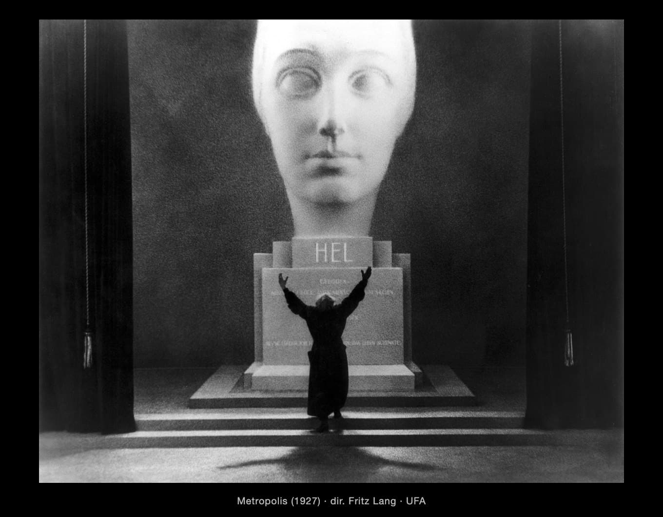 A black-and-white film still from Metropolis (1927). A huge pale stone head of a woman, glowing white, fills the top centre of the frame above a stepped stone plinth carved with the name ‘HEL’. Far below it, a small man in a long black coat stands on the steps with his back to us, arms flung up towards the face. Heavy black curtains hang at both sides like a stage. His long shadow stretches across the floor towards the camera. Caption: ‘Metropolis (1927) · dir. Fritz Lang · UFA’.