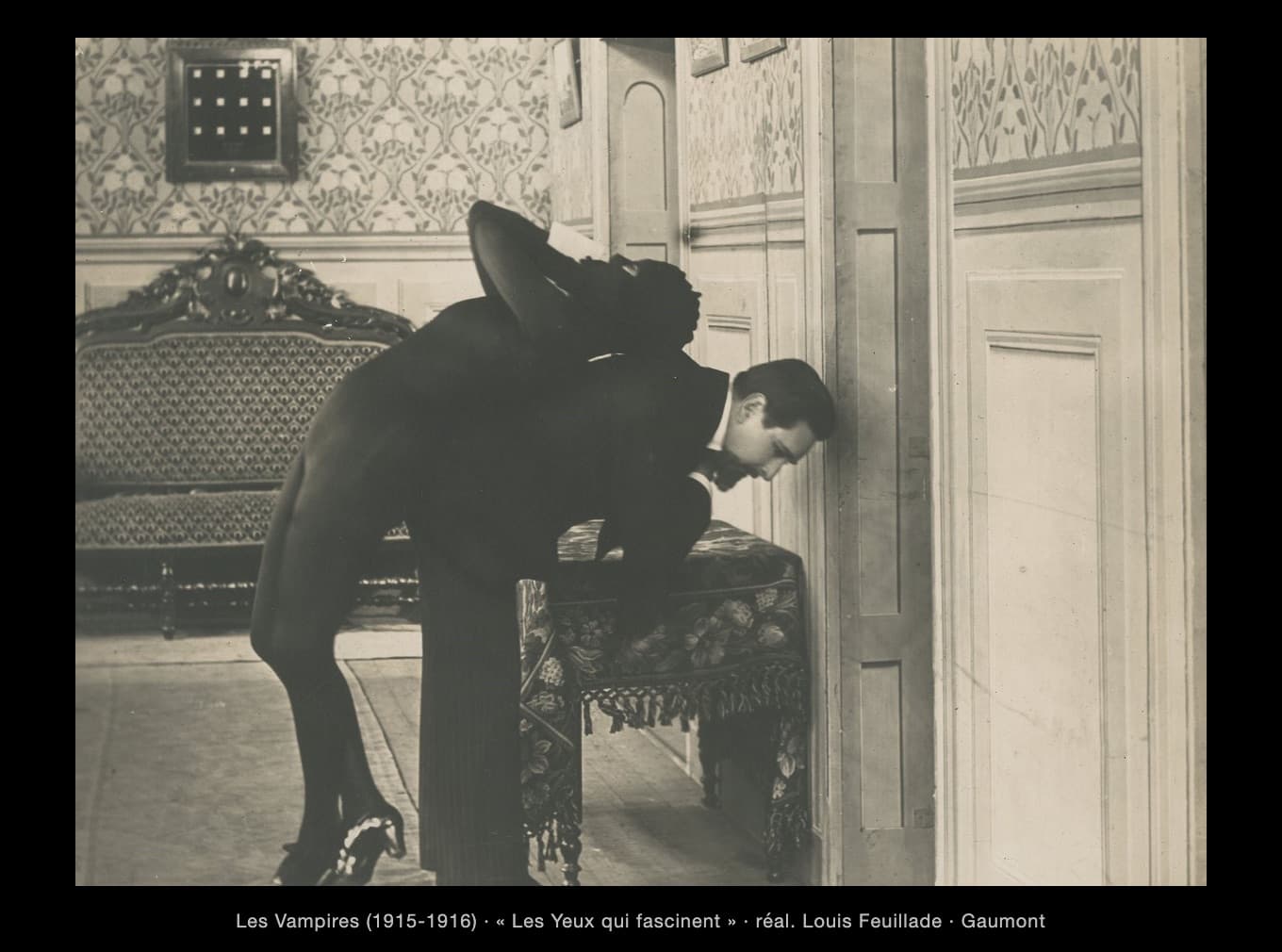Une image en noir et blanc du film muet Les Vampires (1916). Dans un salon bourgeois — papier peint à motifs de fleurs, canapé au dossier sculpté, petite table couverte d’un tapis à franges — un homme en costume noir, penché en avant, porte sur son épaule une femme vêtue d’un maillot noir moulant : sa tête est cachée derrière le dos de l’homme, ses jambes pendent. L’homme colle la tête contre une haute porte claire qui occupe tout le tiers droit de l’image, les yeux baissés, comme s’il écoutait. Les deux corps noirs forment une seule masse sombre sur le décor clair. Légende : « Les Vampires (1915-1916) · “Les Yeux qui fascinent” · réal. Louis Feuillade · Gaumont ».